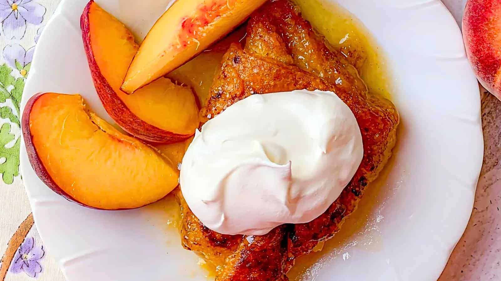 A white plate with a serving of peach cobbler topped with a dollop of whipped cream. Beside the cobbler are several slices of fresh peach. The background includes a partially visible peach and a flowery placemat.