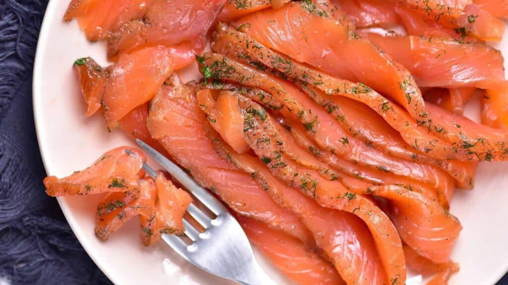 Slices of cured salmon arranged on a white plate with a fork, garnished with herbs.