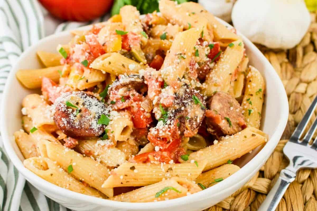 A bowl of penne pasta with sliced sausage, tomatoes, and grated cheese, garnished with herbs. A fork is placed beside the bowl.