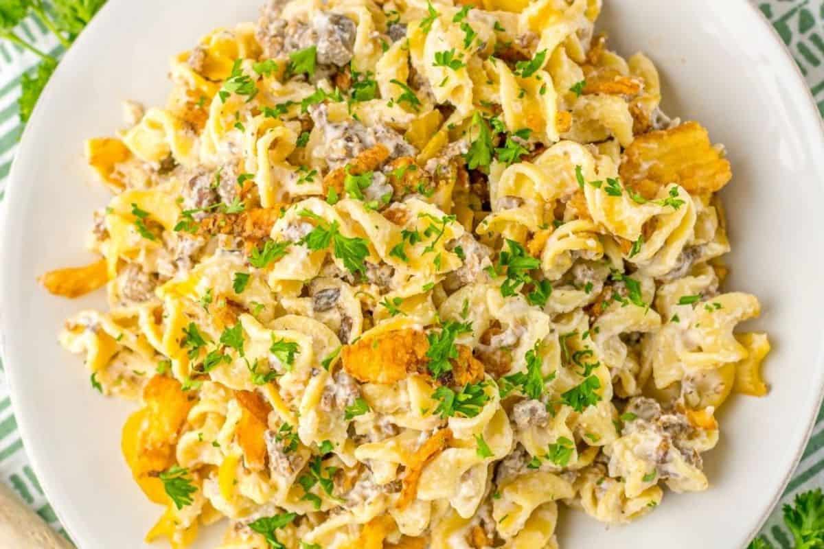 A plate of creamy pasta mixed with ground beef, garnished with chopped parsley and topped with crunchy chips.