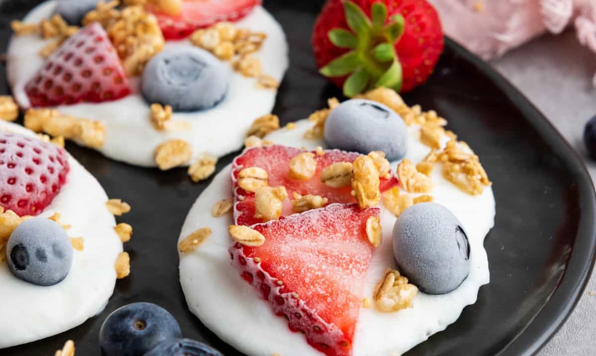 Yogurt rounds topped with sliced strawberries, whole blueberries, and granola are arranged on a black plate.