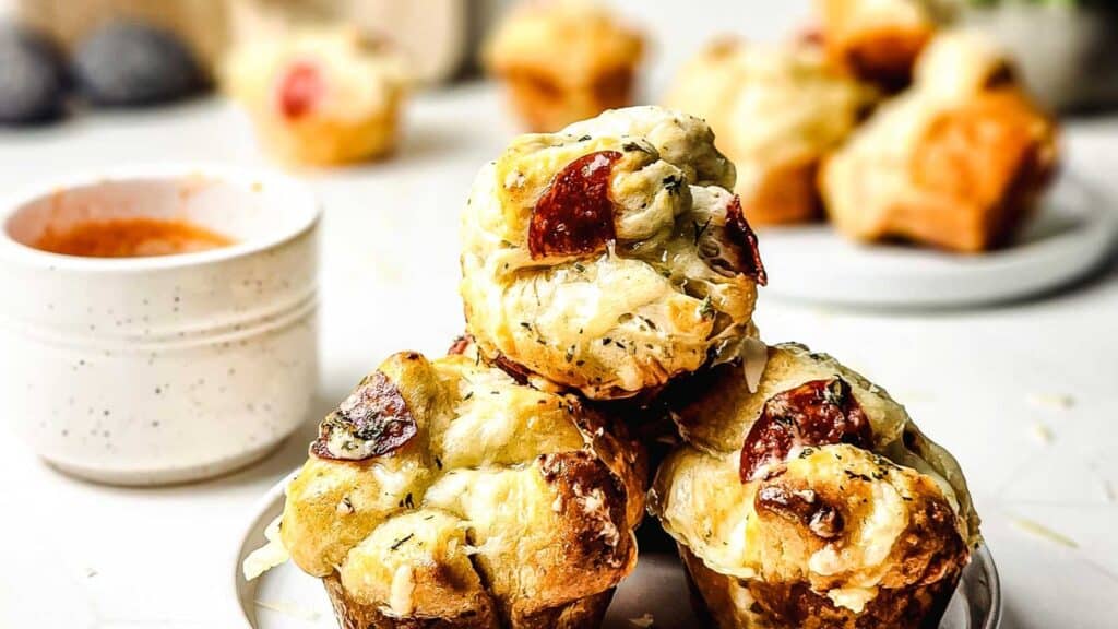 Three savory muffin-like pizza bites with melted cheese and pepperoni are stacked on a plate, with a cup of dipping sauce and more pizza bites in the background.