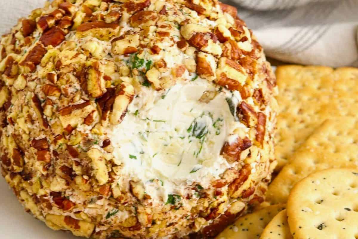 A cheese ball coated with chopped nuts and herbs is served on a plate alongside round crackers.