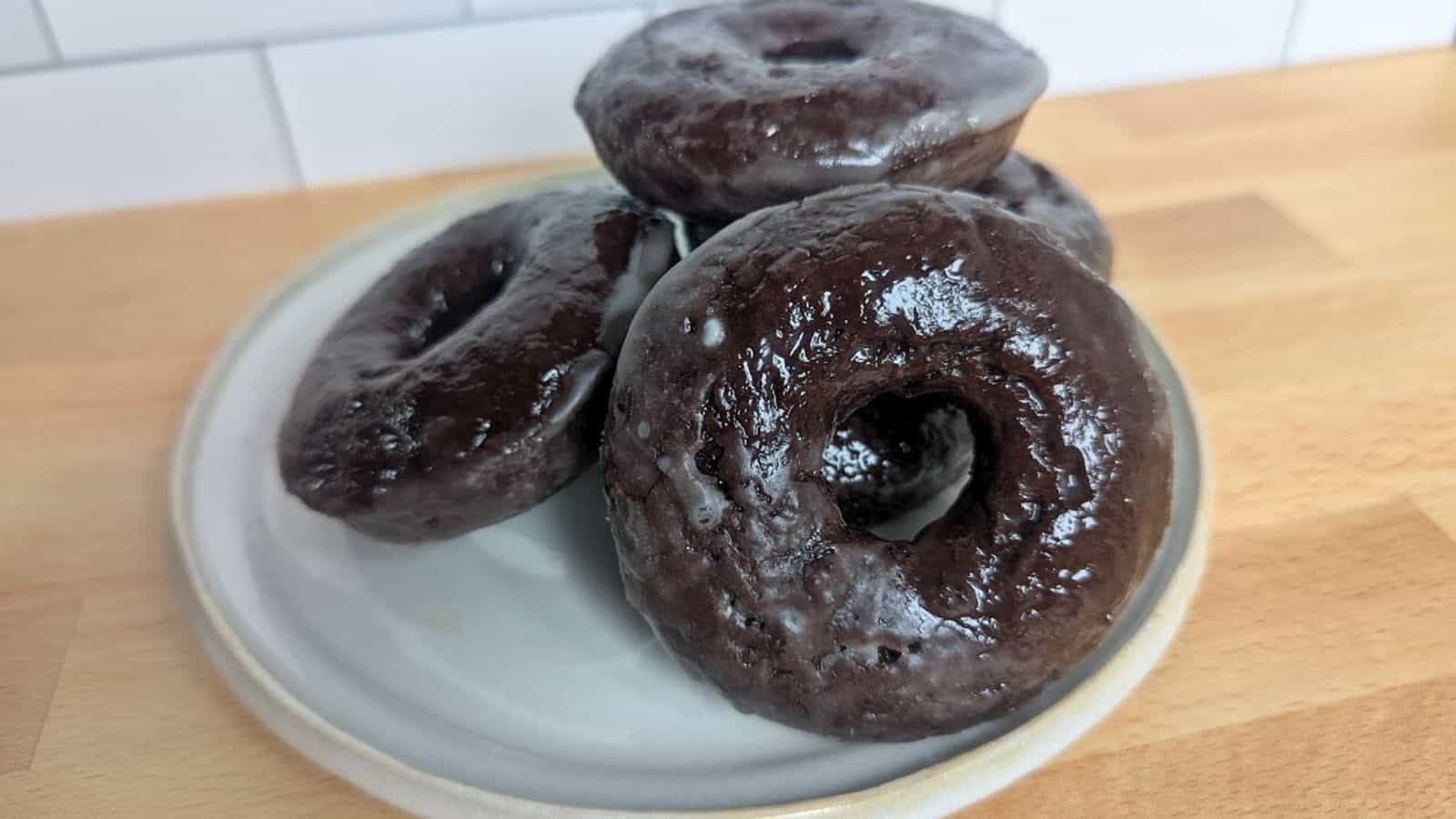 Image shows a  stack of glazed chocolate donuts sits on a white plate atop a wooden surface, with a tiled wall in the background.