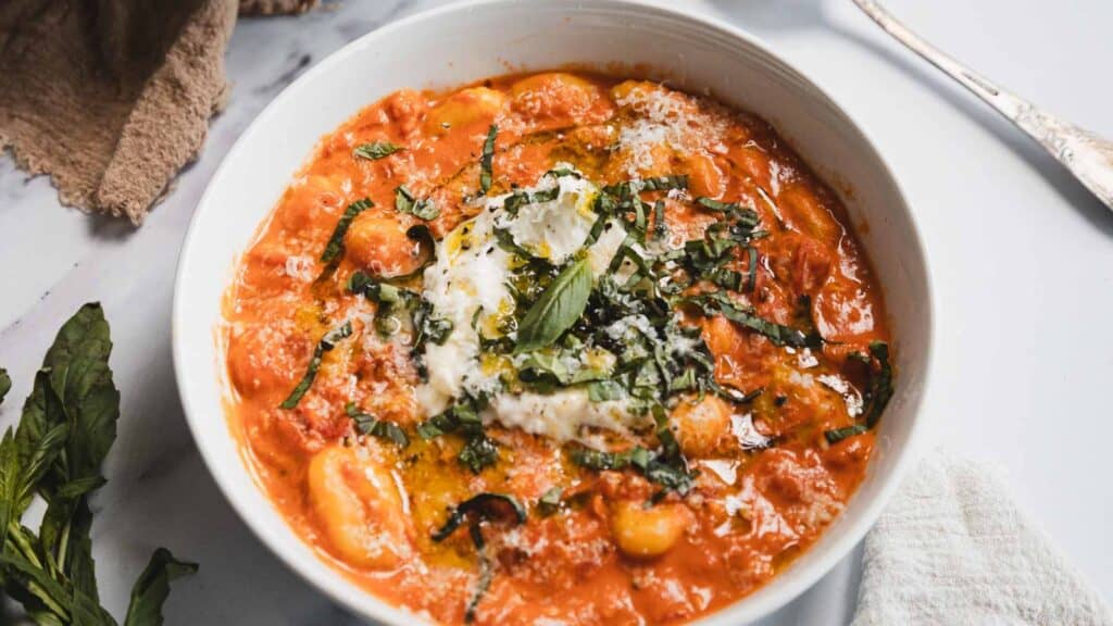 A bowl of gnocchi in tomato sauce topped with cheese, fresh basil, and a drizzle of olive oil sits on a white table with a fork and napkin nearby.