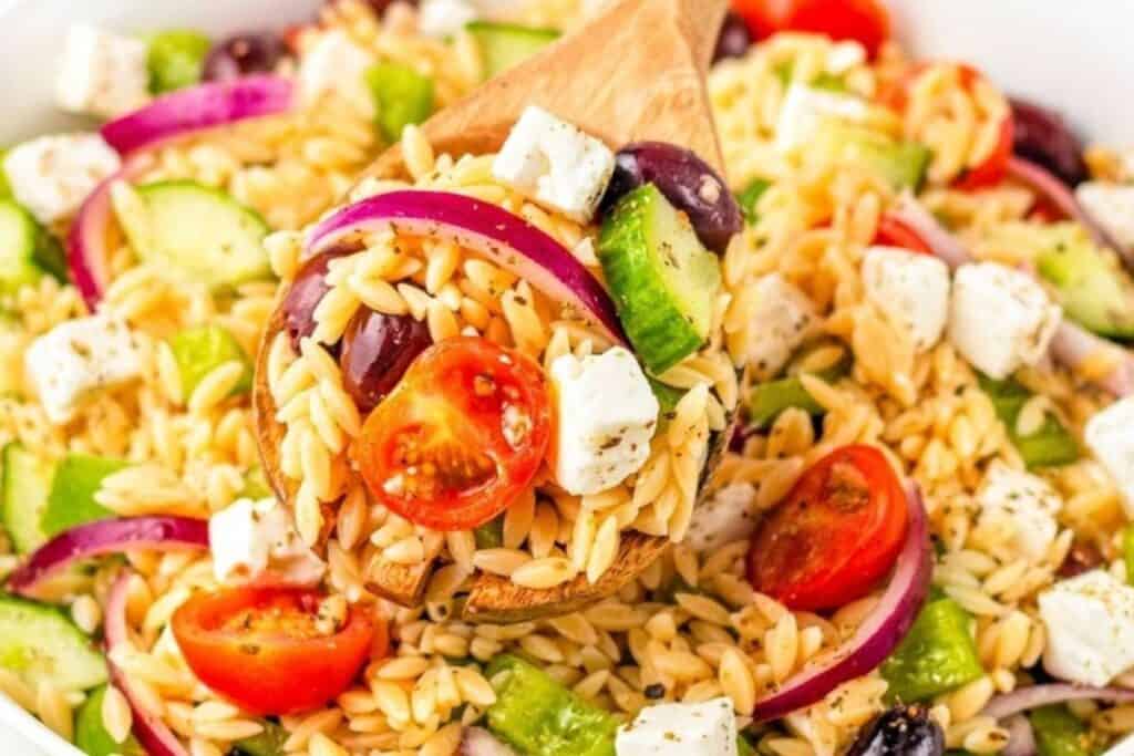 A wooden spoon lifts a serving of orzo pasta salad with cherry tomatoes, cucumber, red onion, feta cheese, and olives from a white bowl.