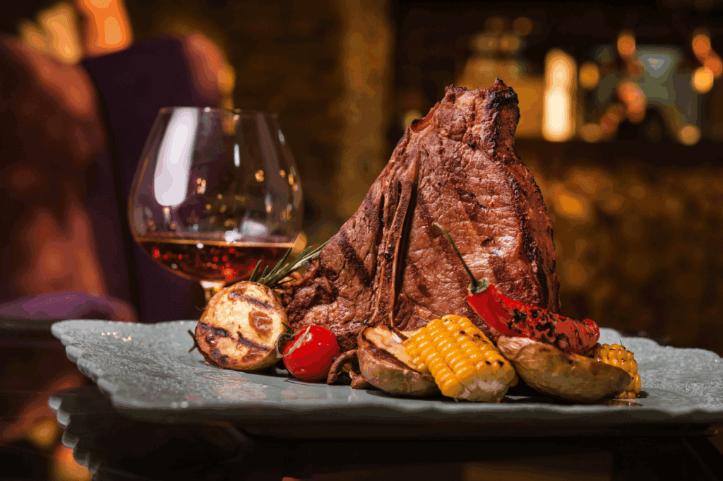 A grilled T-bone steak is served with roasted potatoes, corn, a red pepper, and a cherry tomato, showcasing delicious BBQ flavors and perfect bourbon pairings on a square plate.