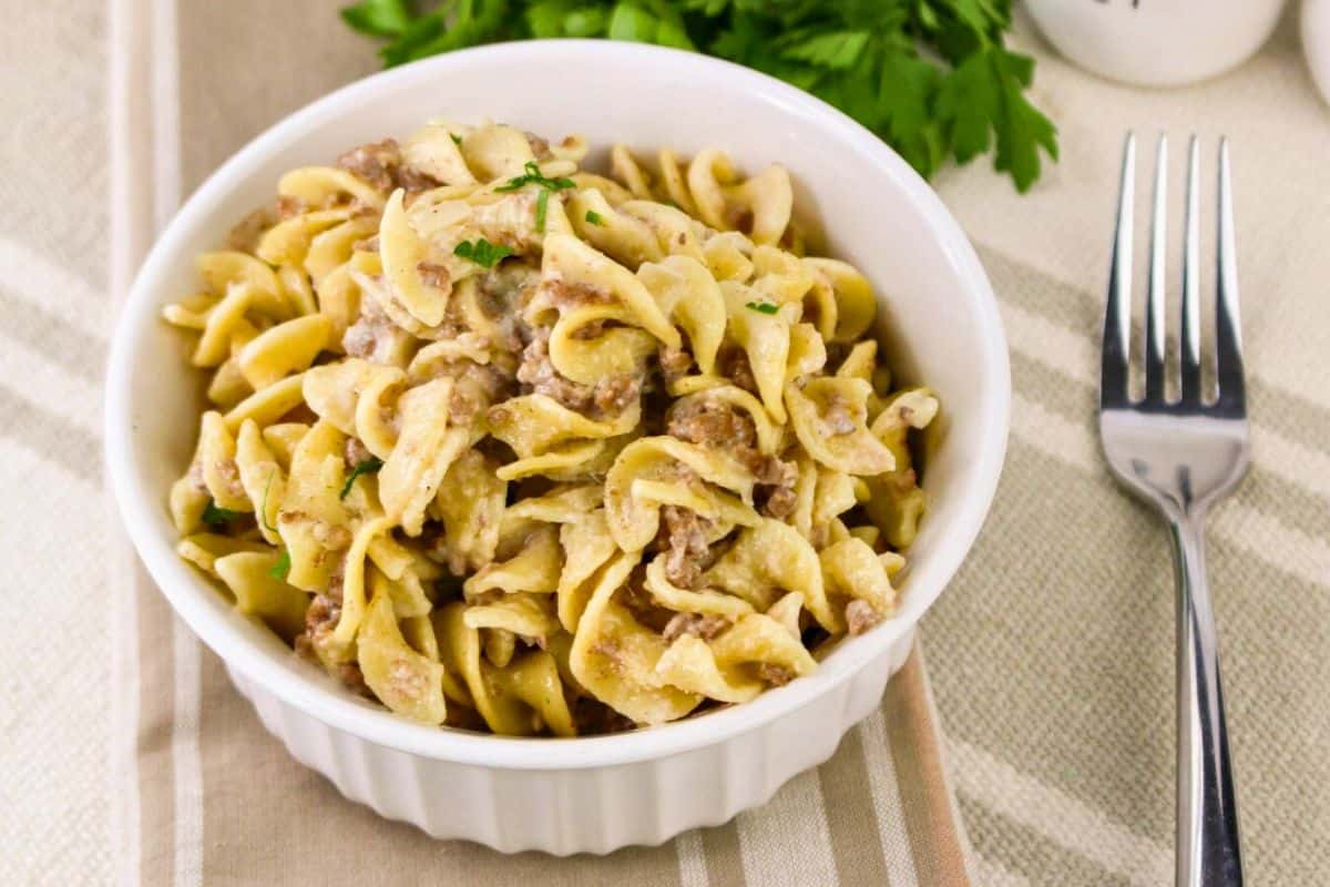 A bowl of creamy beef stroganoff with egg noodles on a striped cloth next to a fork and a sprig of parsley.
