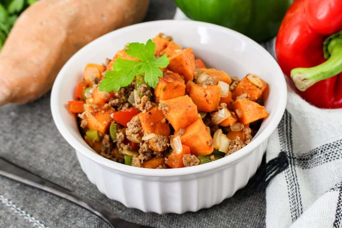 A white bowl filled with a sweet potato and ground beef dish, garnished with a green herb. Surrounding the bowl are a sweet potato, green pepper, and red pepper on a cloth.