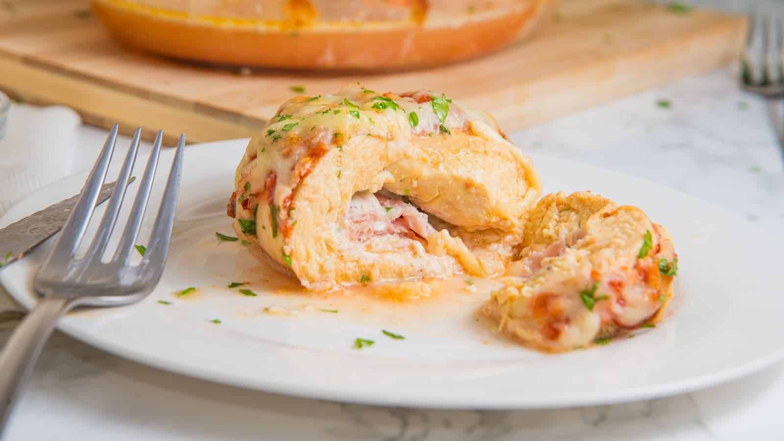 A stuffed chicken breast with melted cheese and herbs on a white plate, partially cut open, with a fork beside it.