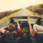 Two people ride in a convertible on an empty road, with one raising their hands. Flowers are on the console, and the scene is sunny with open countryside.