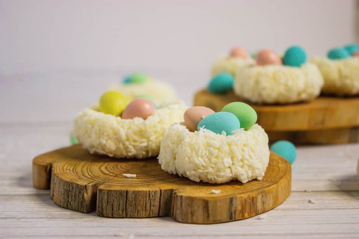 Coconut macaroons shaped like nests, each holding colorful candy eggs, create a charming display on a wooden board and white surface.