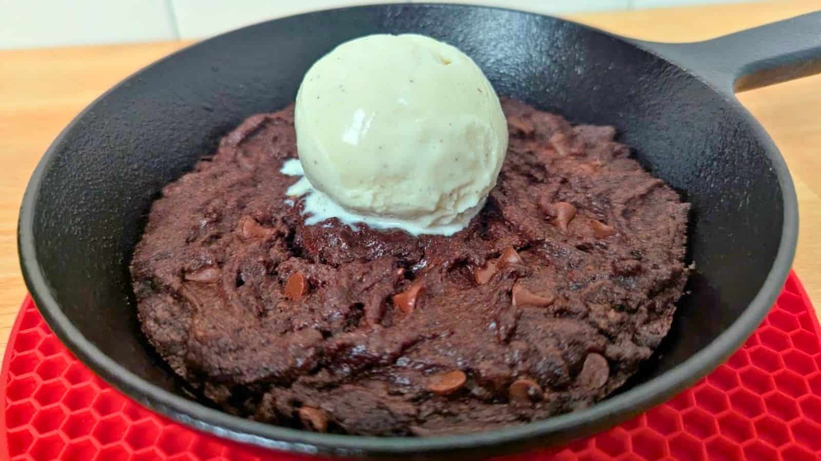 Image shows a chocolate chip skillet cookie topped with a scoop of vanilla ice cream, served in a black pan on a red mat.