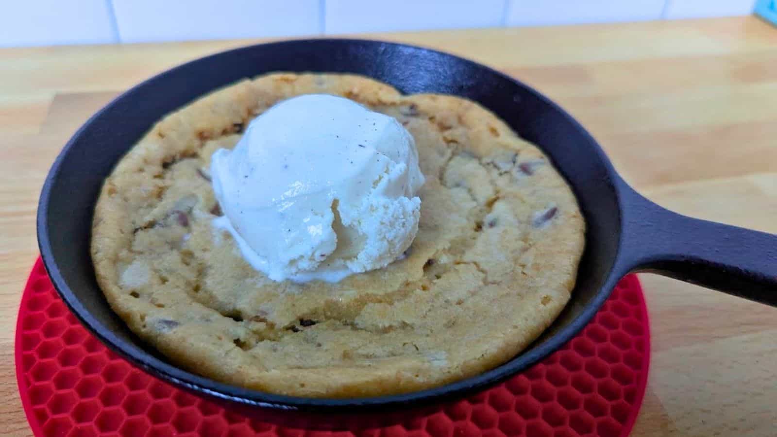Image shows a chocolate chip cookie baked in a small cast iron skillet, topped with a scoop of vanilla ice cream, sits on a red silicone trivet on a wooden surface.
