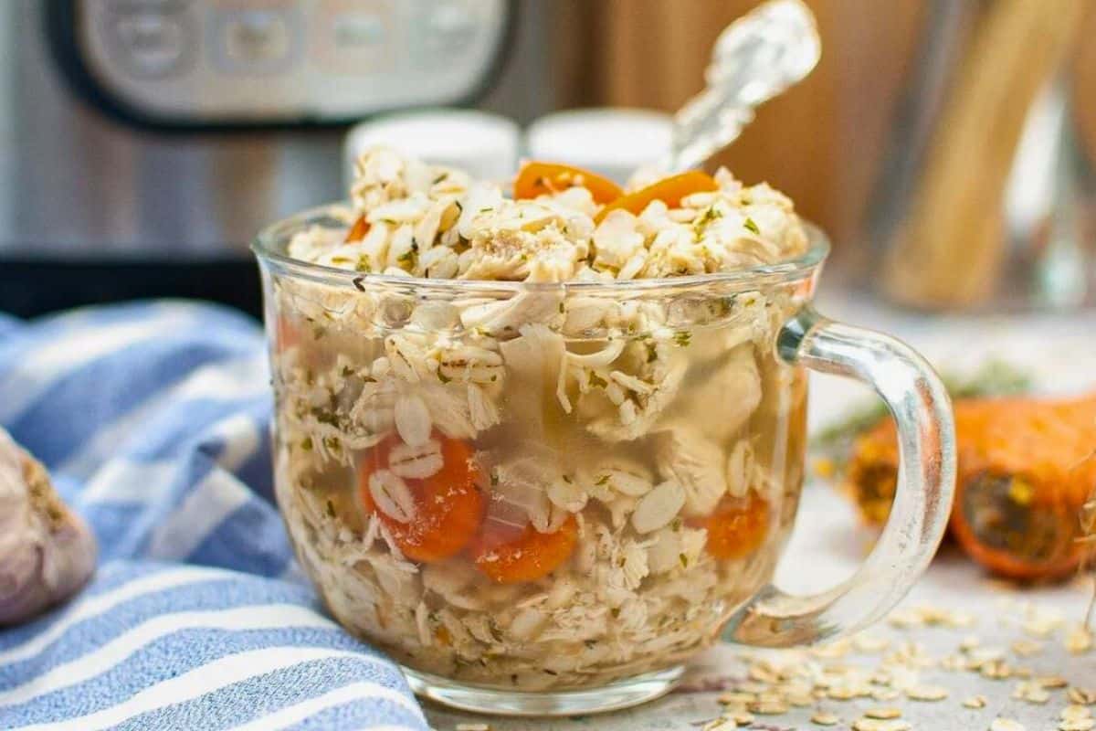 A clear glass mug filled with chicken soup, oats, and carrot slices sits on a countertop with a silver spoon inside.
