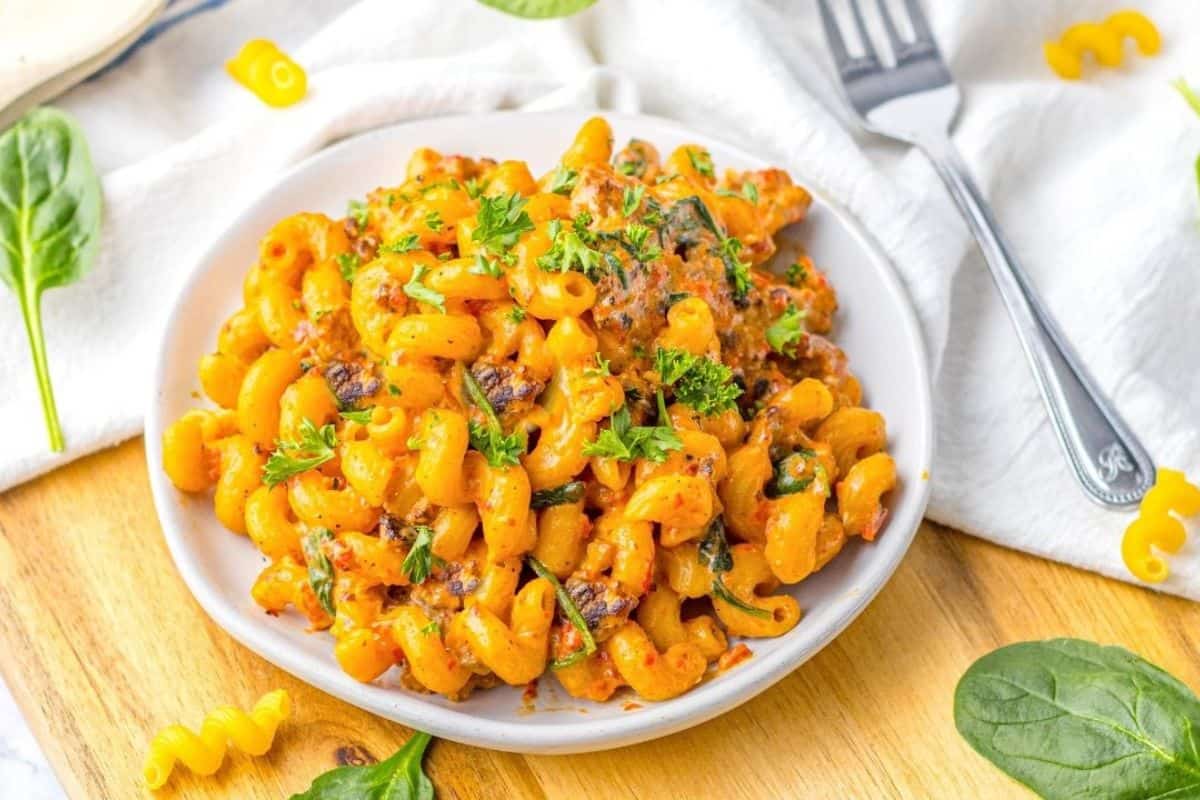 A plate of creamy macaroni pasta with herbs and vegetables, garnished with chopped parsley, on a wooden board with spinach leaves nearby.