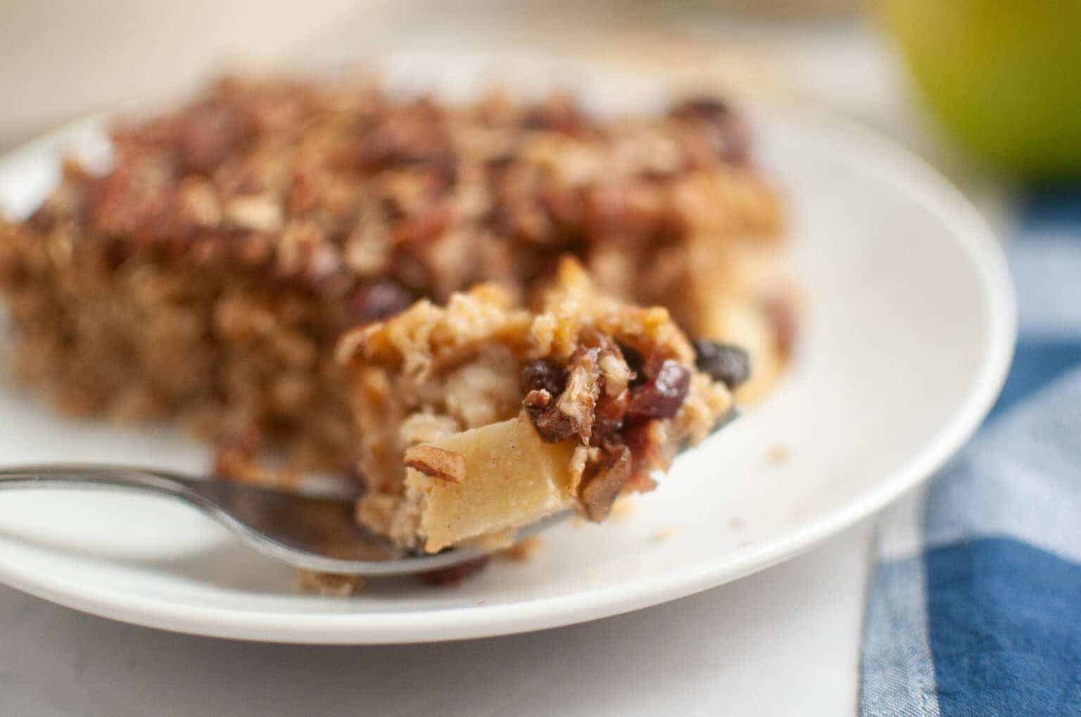 A close-up of a fork holding a bite of Apple-Pecan Baked Oatmeal. on a white plate, with the rest of the serving in the background.