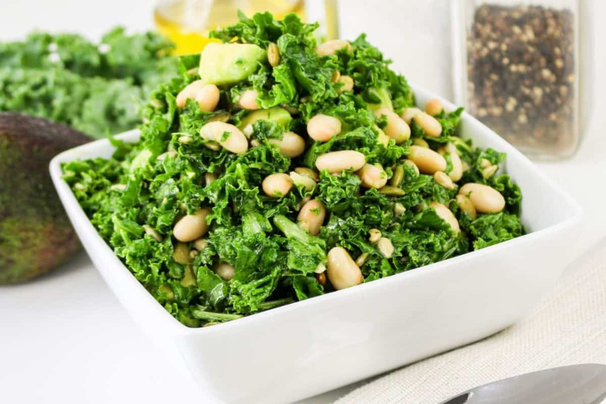 A square white bowl filled with kale salad, featuring beans and avocado pieces, placed on a table with a spoon beside it.