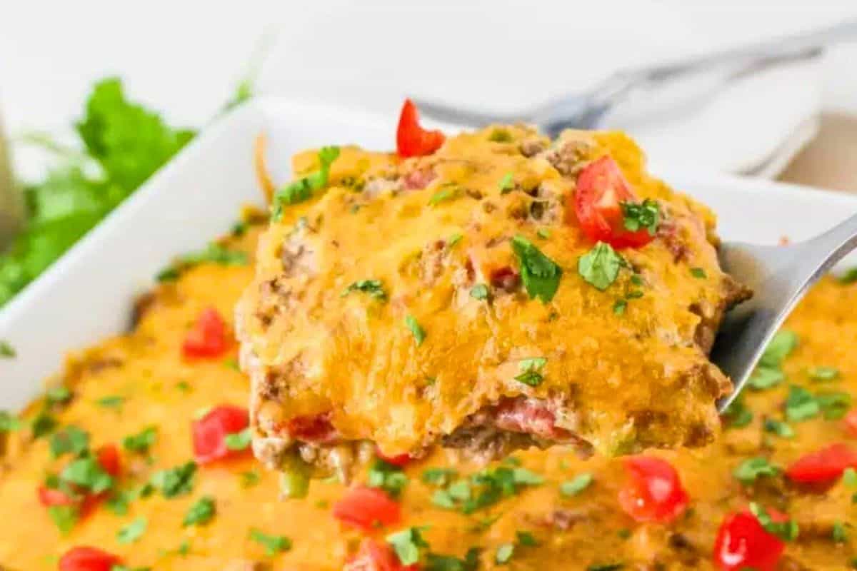 Close-up of a spoon lifting a portion of cheesy beef casserole garnished with chopped tomatoes and herbs from a white baking dish.