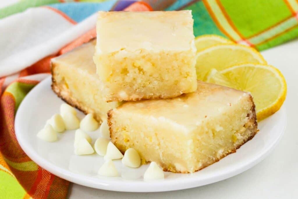 Three lemon brownies stacked on a white plate with white chocolate chips and lemon slices, set against a colorful cloth background.