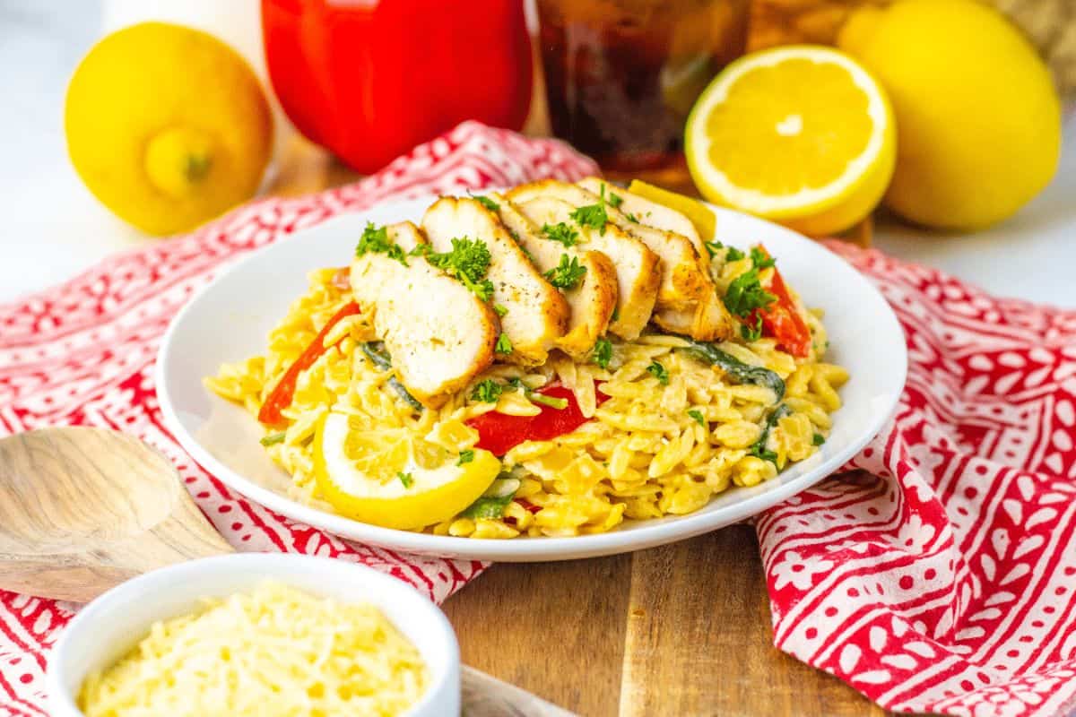 A plate of orzo pasta topped with sliced grilled chicken, garnished with parsley and lemon. Nearby are bell peppers, lemons, and a bowl of grated cheese on a red patterned cloth.