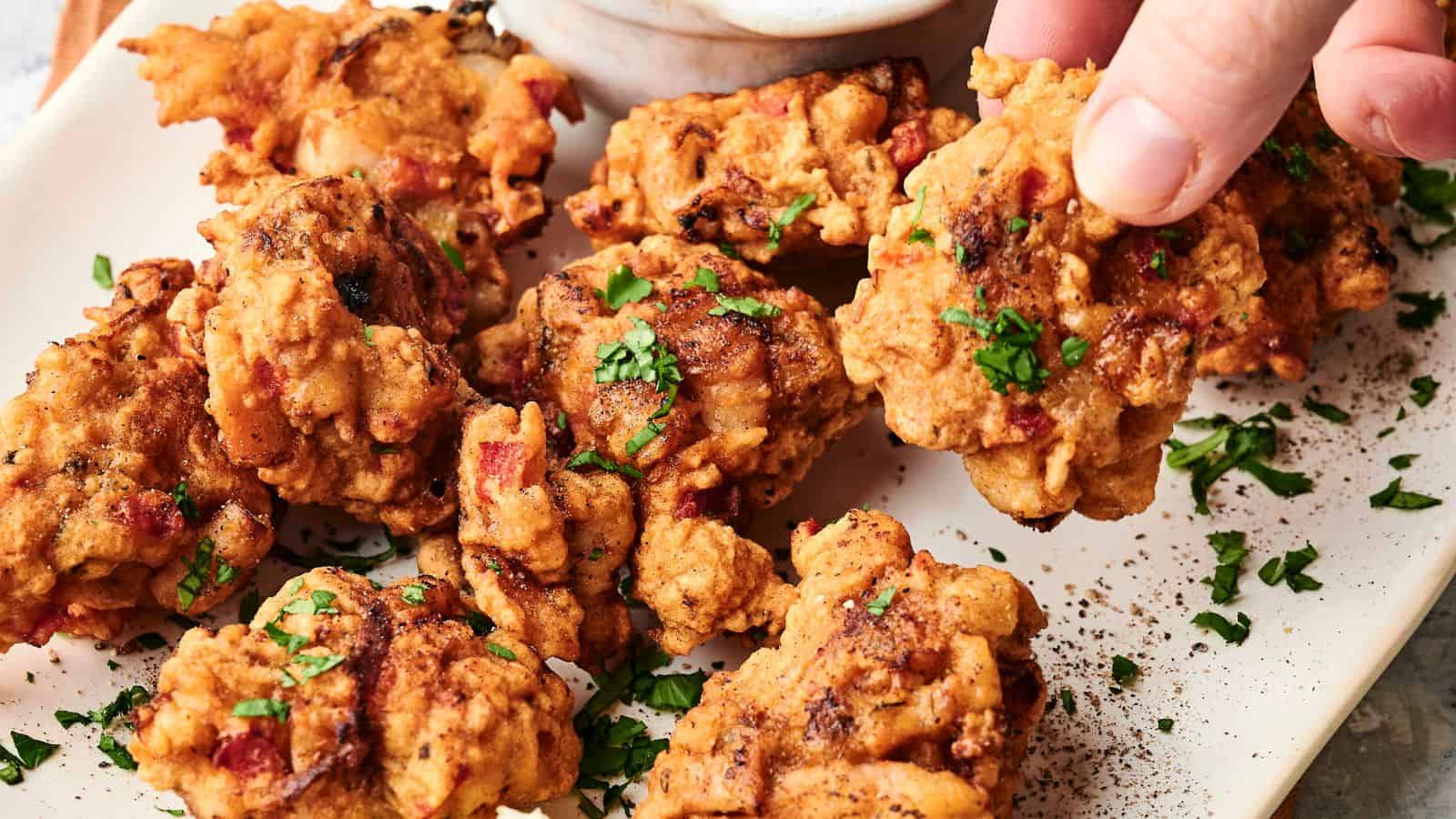 A hand reaches for a piece of crispy, golden-brown fried chicken garnished with chopped parsley on a white plate.