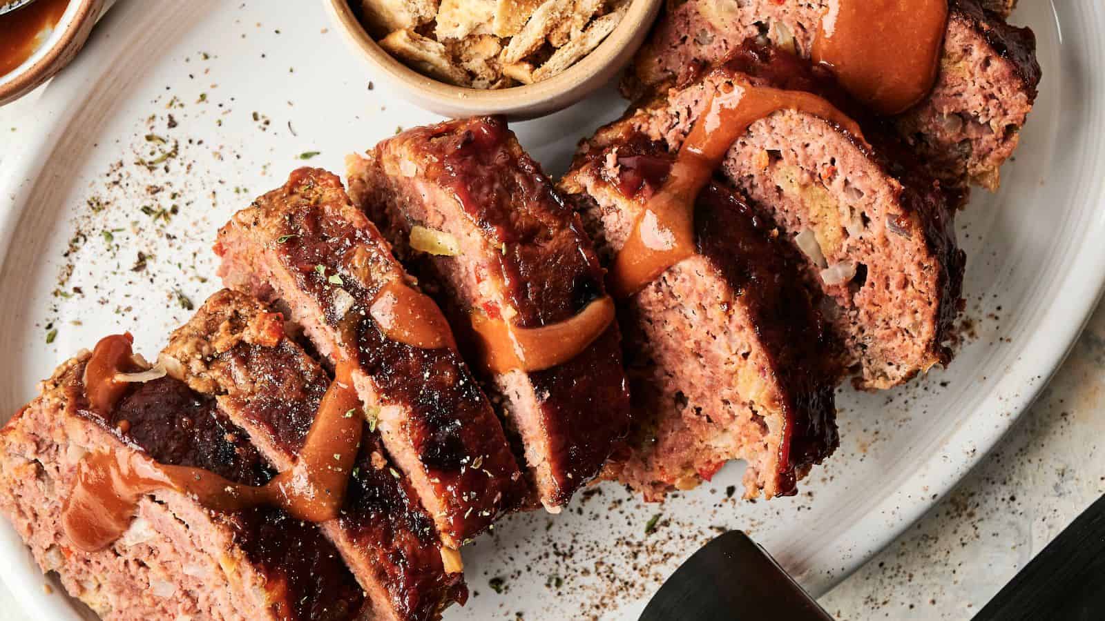 Sliced meatloaf topped with sauce is arranged on a white plate next to a small bowl of crispy toppings.