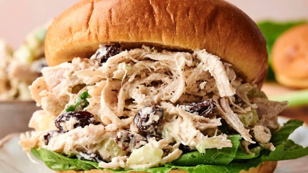 A close-up of a chicken salad sandwich on a bun, with shredded chicken, raisins, celery, and lettuce visible.