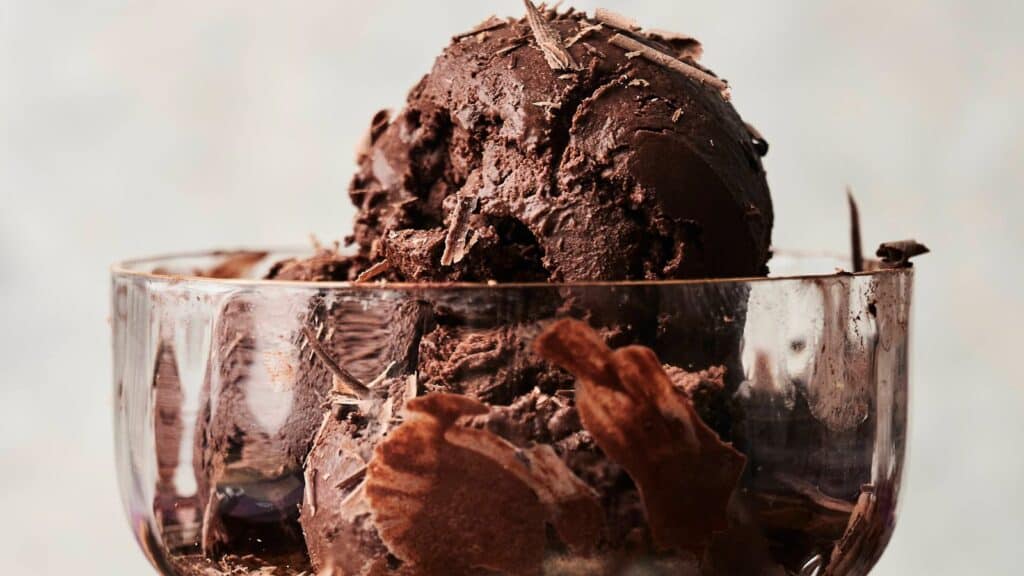 A close-up of two scoops of dark chocolate ice cream with chocolate shavings in a clear glass dessert bowl.