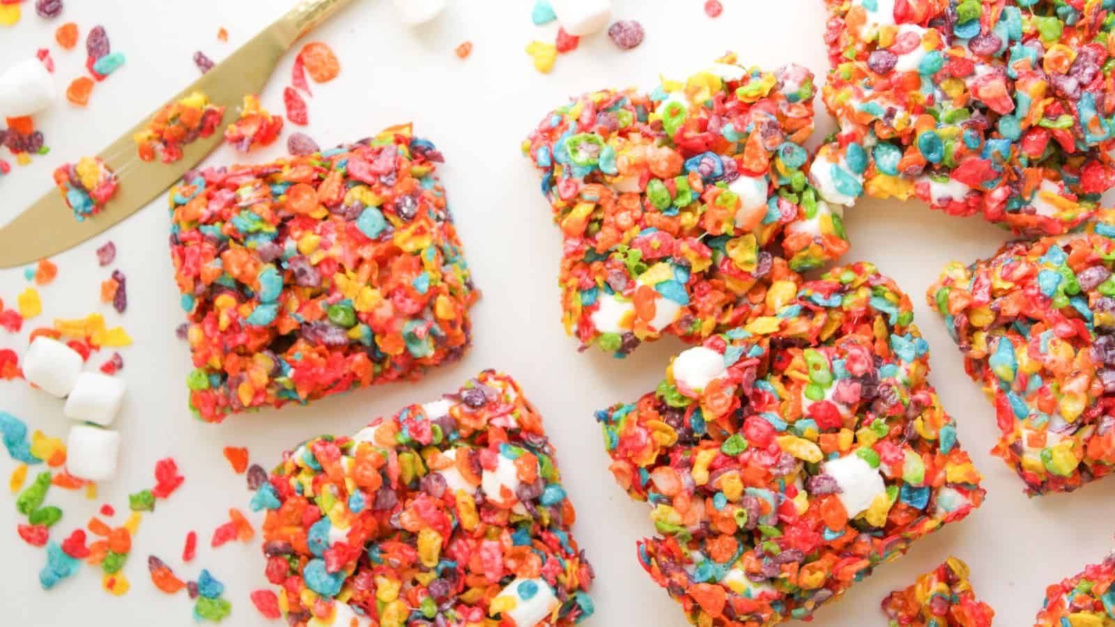 Several square treats made from colorful cereal and mini marshmallows are arranged on a white surface with a butter knife and scattered cereal and marshmallows nearby.