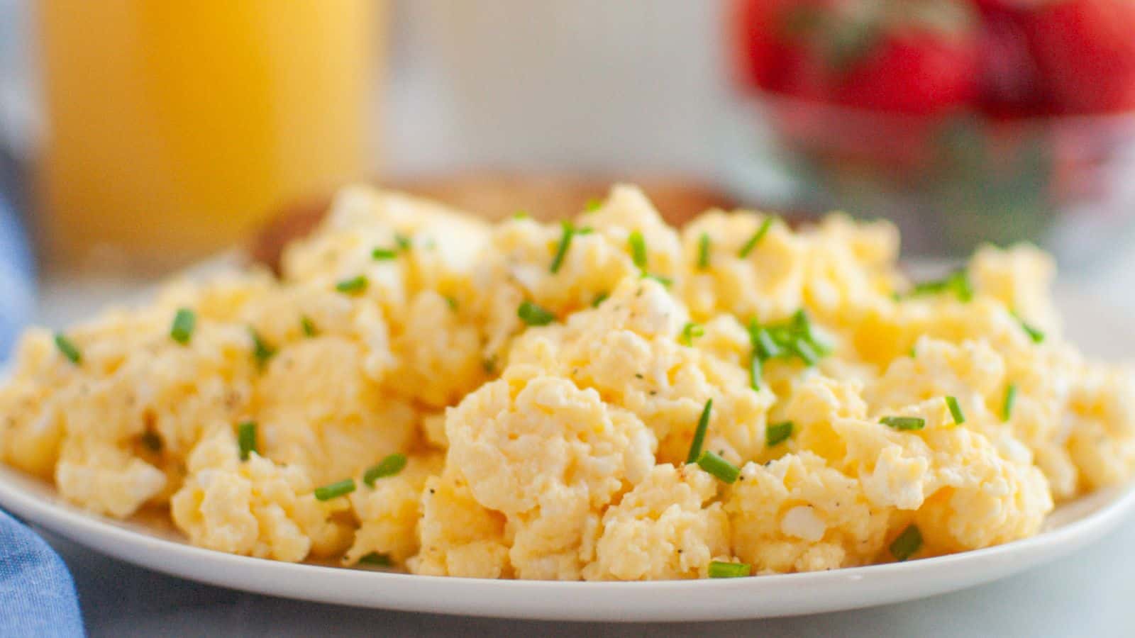A plate of scrambled eggs garnished with chopped chives, with a blurred background showing a bowl of strawberries and a glass of orange juice.