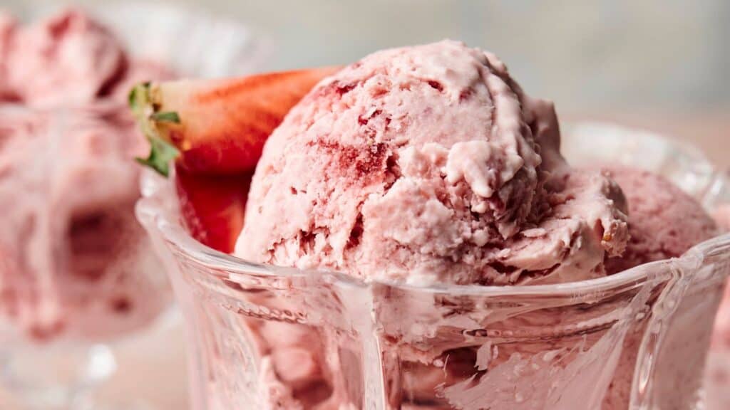 Close-up of strawberry ice cream scoops in a glass dish, garnished with a fresh strawberry.