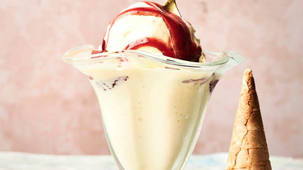 A glass dish filled with vanilla ice cream topped with red syrup, alongside an empty ice cream cone on the right.