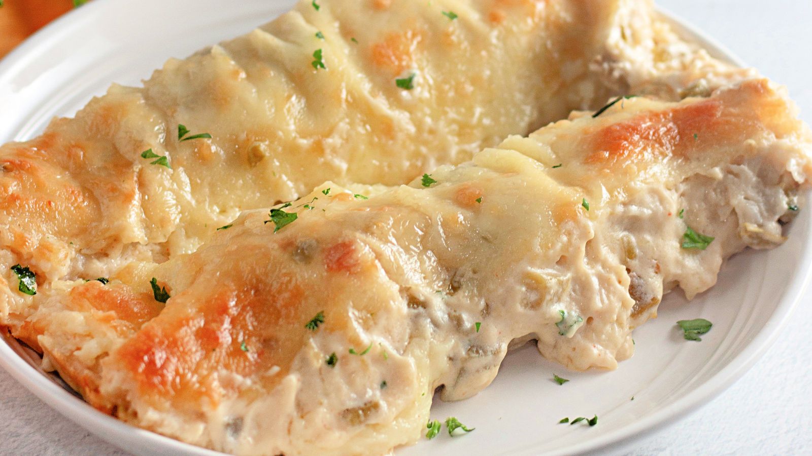 Two White Chicken Enchiladas filled with creamy sauce and topped with melted cheese, served on a white plate with a garnish of chopped parsley.