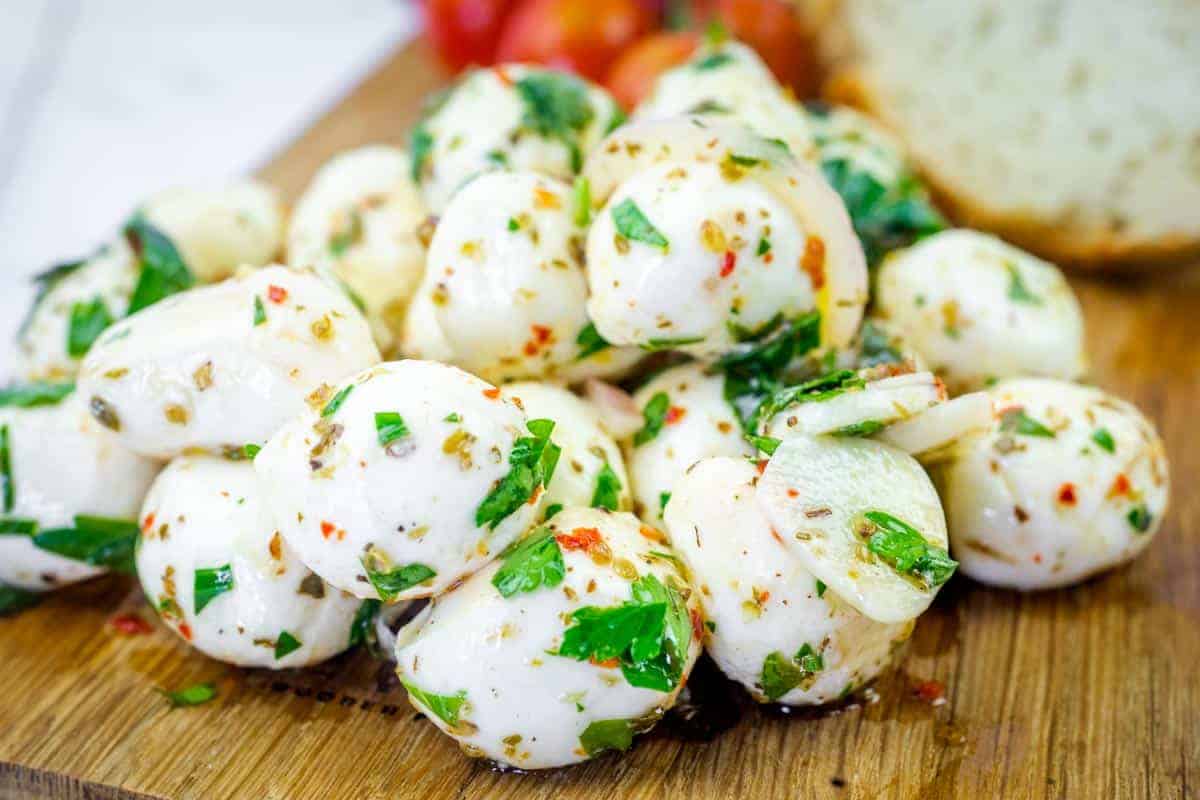 A pile of keto marinated mozzarella balls mixed with chopped herbs and spices on a wooden board, with bread and tomatoes in the background.