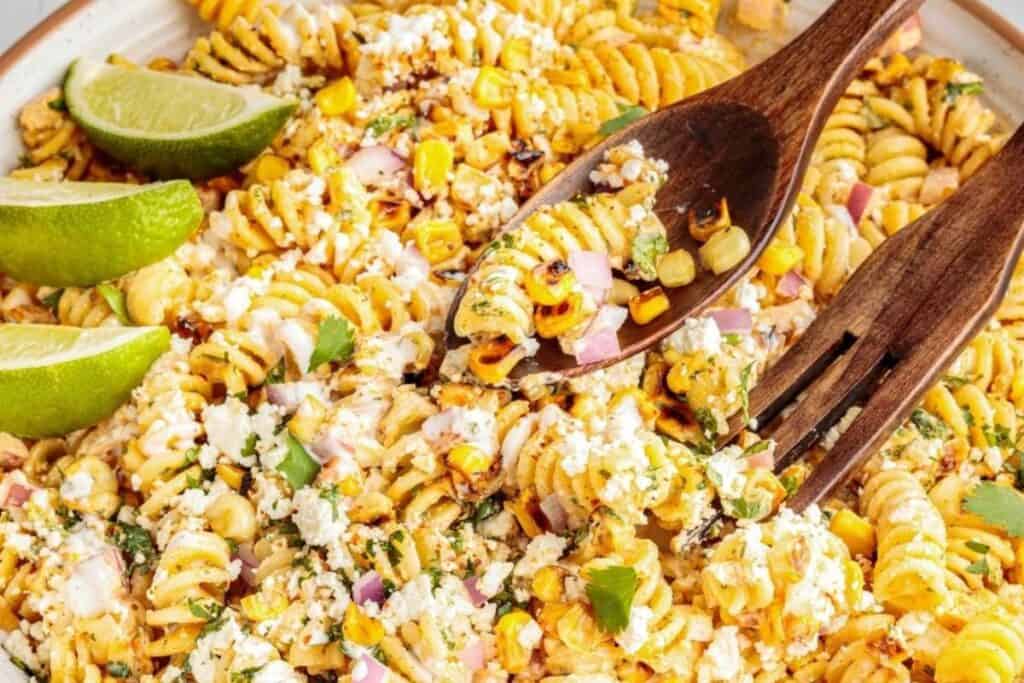 A bowl of rotini pasta salad mixed with corn, cotija cheese, red onion, cilantro, and lime wedges, served with wooden utensils.