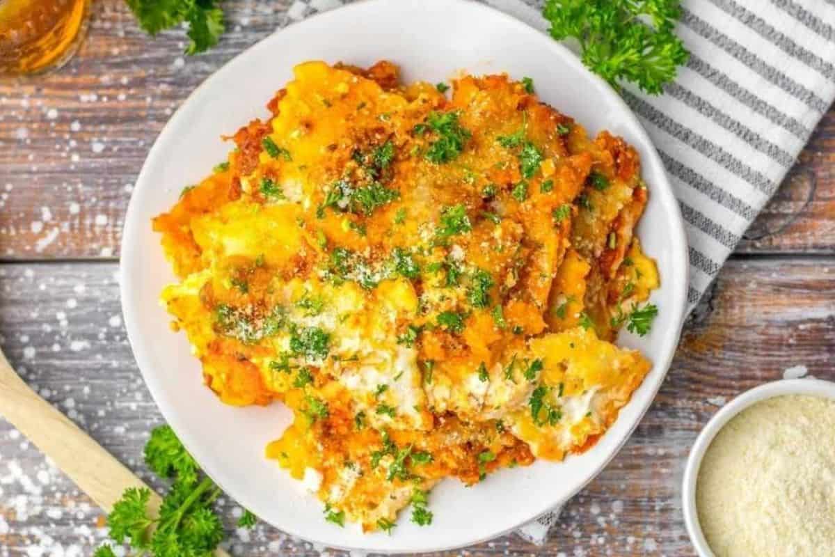 A plate of baked lasagna topped with grated cheese and fresh parsley, placed on a wooden table beside a bowl of grated cheese and a striped napkin.