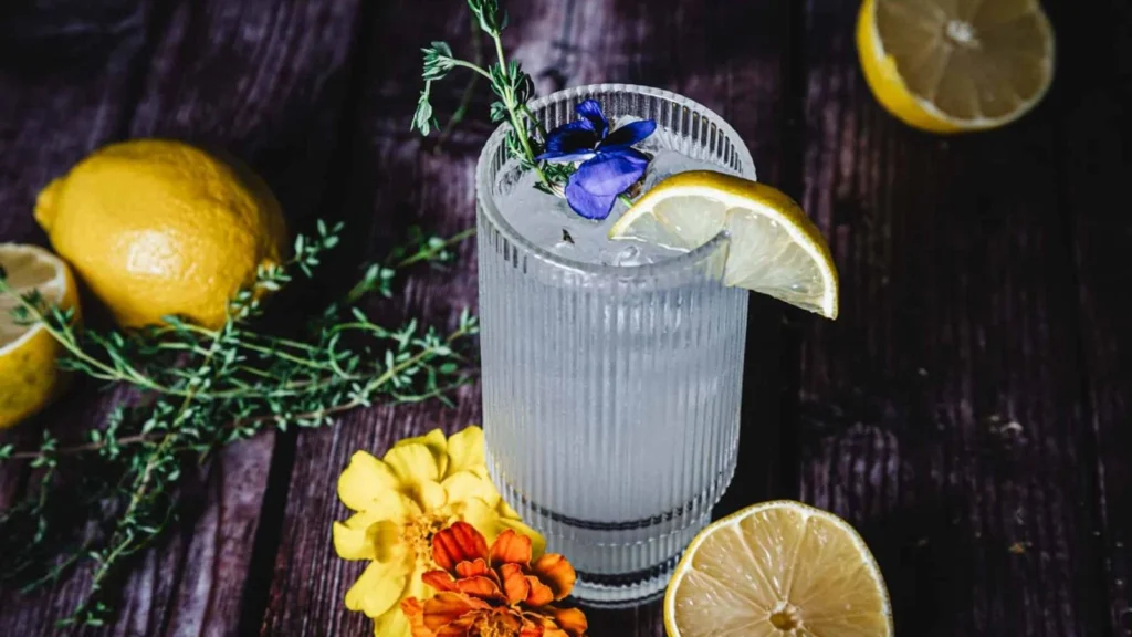 A clear cocktail garnished with a lemon slice, blue flower, and herbs in a ribbed glass, surrounded by lemons and flowers on a dark wooden surface.