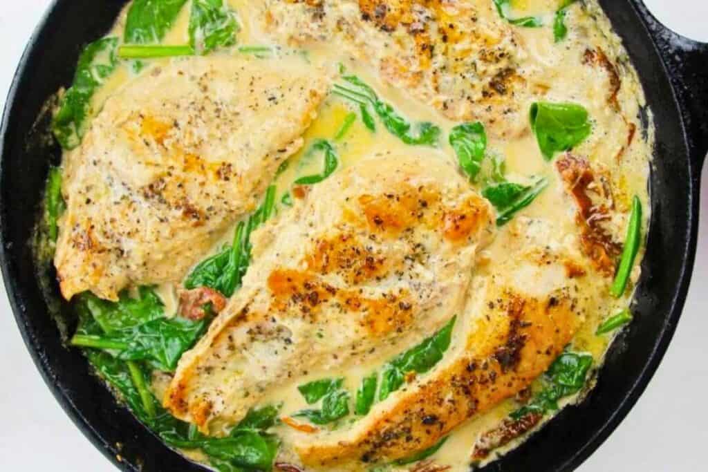 Three seasoned chicken breasts simmer in a creamy sauce with wilted spinach and sun-dried tomatoes in a black skillet.