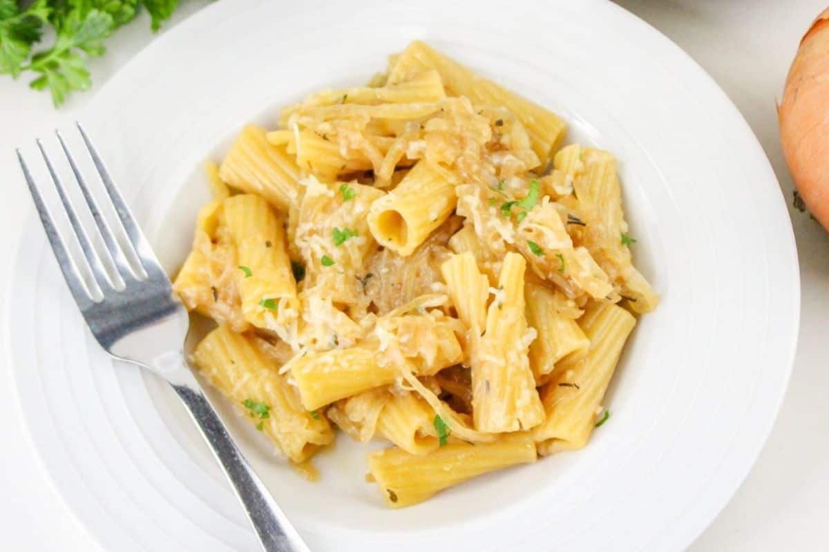 A plate of rigatoni pasta with shredded cheese and herbs, next to a fork on a white dish. An onion and parsley are in the background.