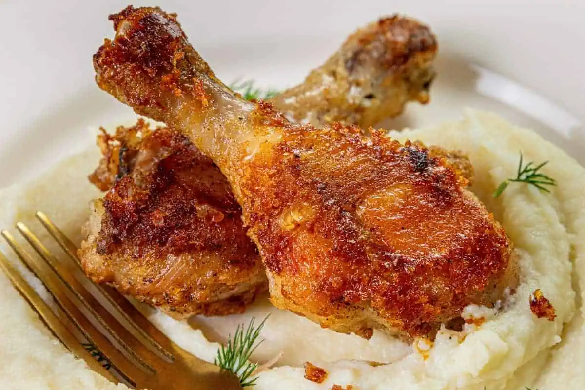 Two crispy fried chicken drumsticks on a bed of creamy mashed potatoes, garnished with dill. A fork is placed beside the dish. The chicken is golden brown and the potatoes have a smooth texture.