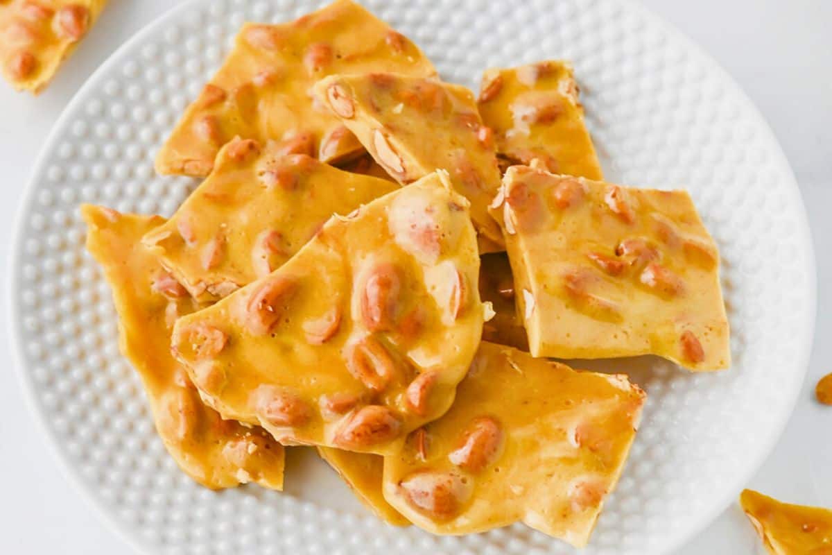 A white plate holds several pieces of golden brown peanut brittle with visible peanuts embedded in the candy.
