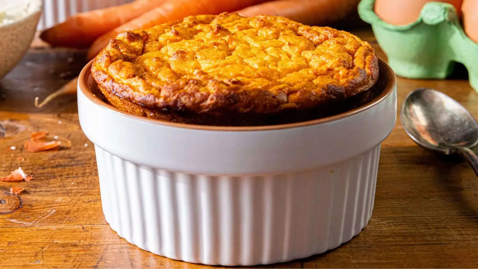 A golden-brown carrot souffl&eacute; in a white ramekin sits on a wooden table. Nearby are scattered ingredients: a spoon, eggs in a green carton, and aged sharp cheddar, embodying the essence of a classic Southern side dish.
