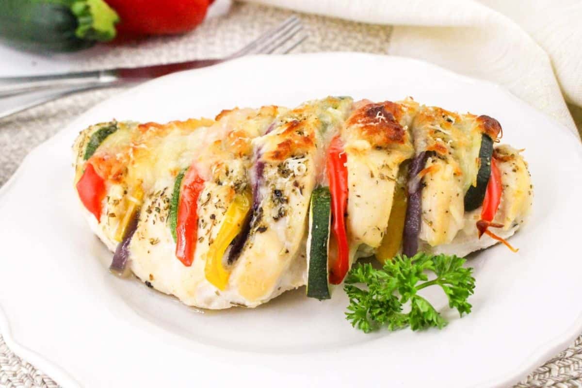 A baked chicken breast stuffed with slices of zucchini, red bell pepper, and cheese, garnished with herbs, on a white plate with a sprig of parsley.