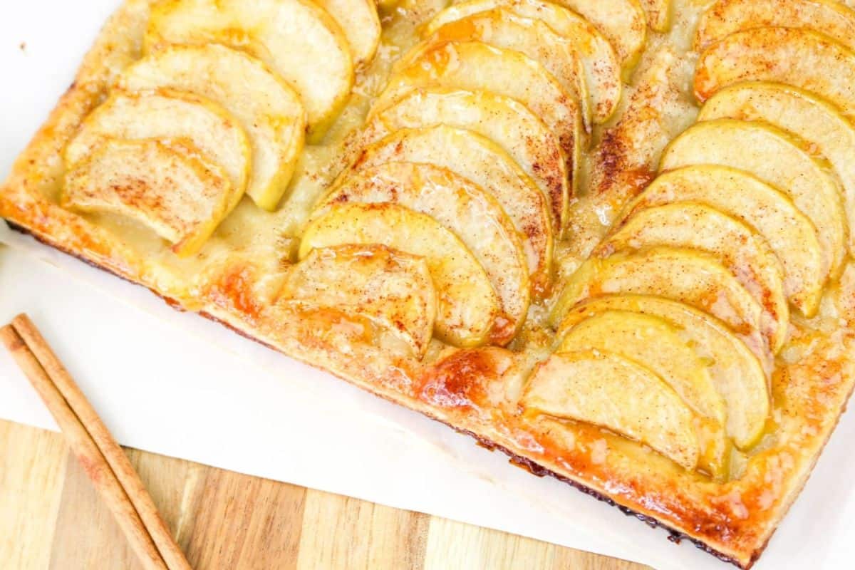 Rectangular apple tart with golden apple slices, a flaky crust, and a light glaze. Two cinnamon sticks lie nearby on a wooden surface.