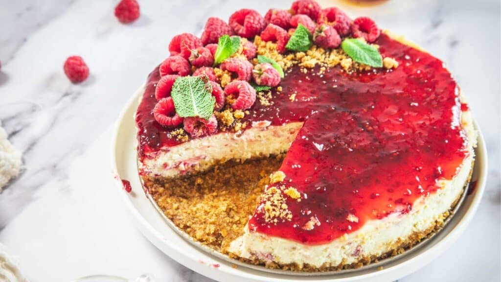 A raspberry cheesecake with a graham cracker crust, topped with raspberry sauce, fresh raspberries, and mint leaves; one slice is missing.