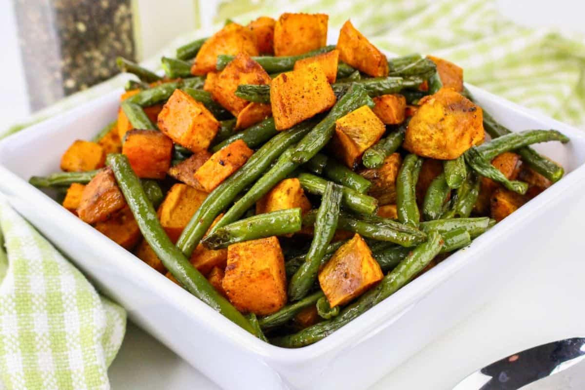 A square white bowl filled with roasted sweet potatoes and green beans, placed on a green checkered cloth, with a spoon and pepper mill visible in the background.
