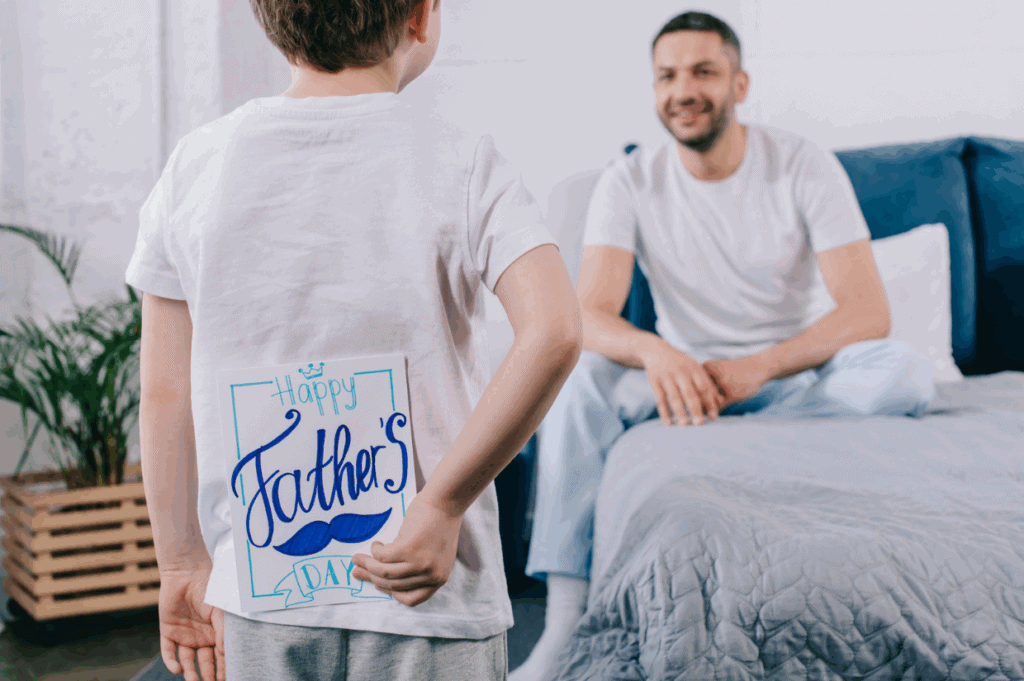 A child hides a "Happy Father's Day" card behind their back while standing near a man sitting on a bed, ready to surprise him on Father's Day.