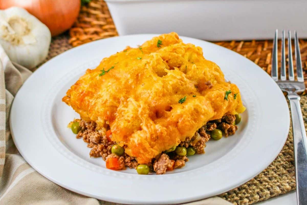 A plate of shepherds pie topped with melted cheese, showing layers of ground meat, peas, and carrots. Near the plate are a fork, garlic, onion, and a striped napkin.