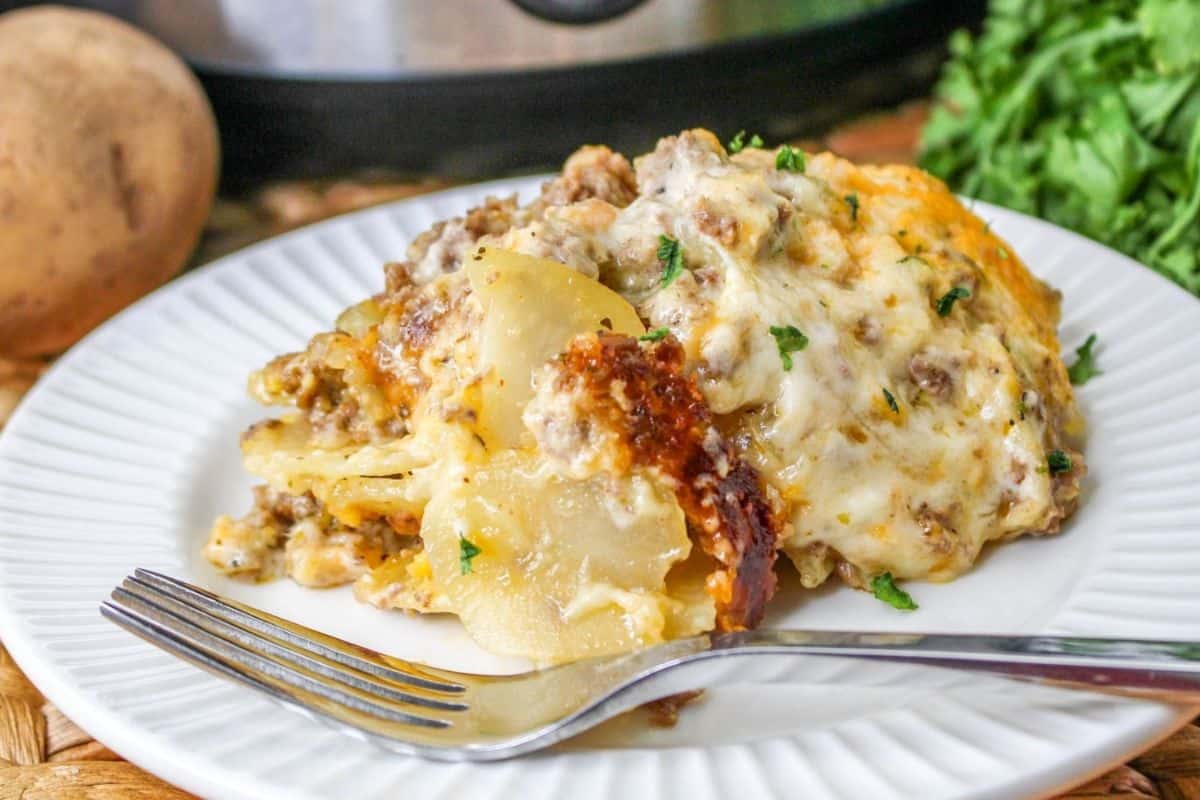 A plate of cheesy beef and potato casserole garnished with herbs, served with a fork. A potato, greenery, and a slow cooker are visible in the background.